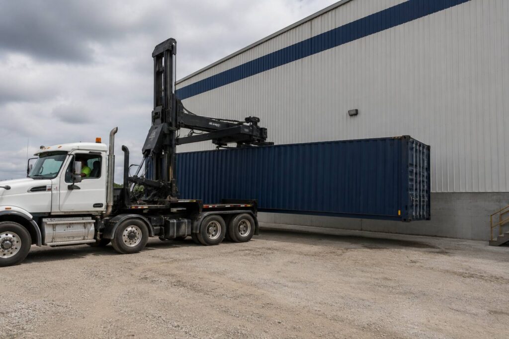 Specialized truck with a side loader handling a navy shipping container at a warehouse